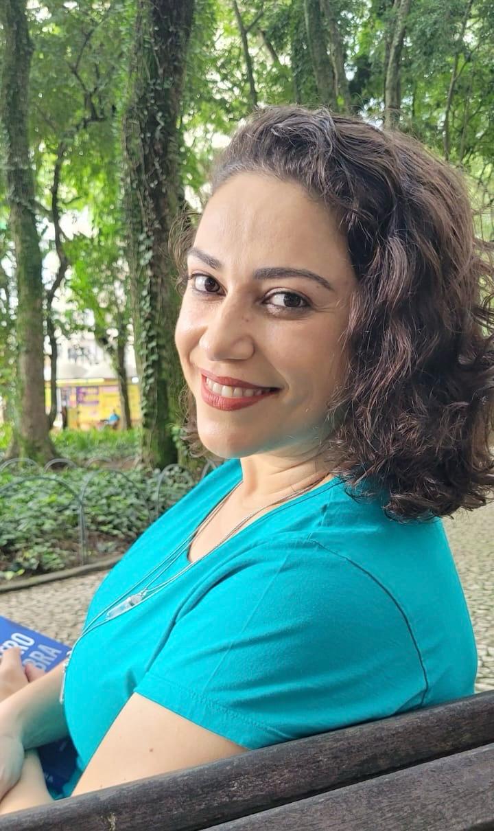 Letícia Máia sorrindo, com blusa azul-turquesa, em uma área arborizada